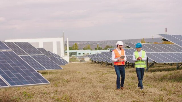 At modern solar panels farm walking two industrial mechanics in a protective equipments with helmets and discussing about some information concept of electrical innovation nature environment