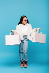full length of shocked preteen girl in pink sunglasses and faux fur jacket holding shopping bags on blue.