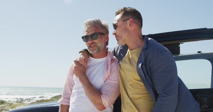 Happy caucasian gay male couple wearing sunglasses embracing by car on sunny day at the beach