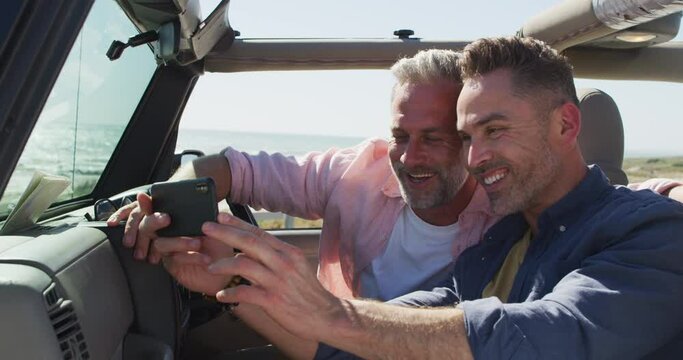 Happy caucasian gay male couple taking selfies sitting in car smiling on sunny day at the beach