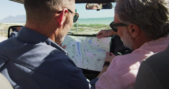 Happy caucasian gay male couple sitting in car reading map and pointing on sunny day at the beach