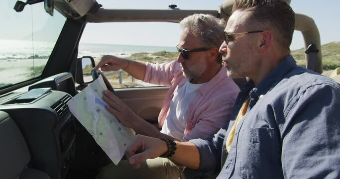 Happy caucasian gay male couple sitting in car reading map and pointing on sunny day at the beach