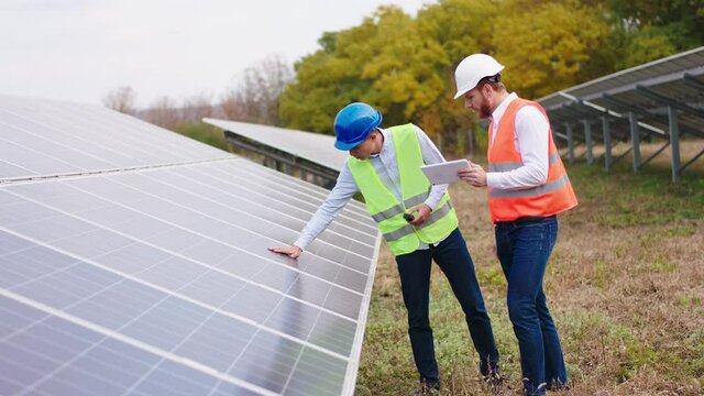 Two ecological engineers at the solar panels park controlling the photovoltaic batteries and using digital tablet to write information they wearing protective helmets concept of electrical innovation