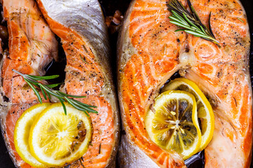 Top view of fresh roasted red salmon fish steaks with herbs, spices and lemon on black metal plate background in the kitchen.