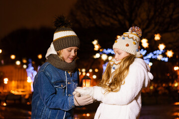 Funny couple of children, girl and boy, hold lantern in their hands against background of christmas lights outside, in warm winter clothes and knitted hats. Christmas and childhood concept, outdoor