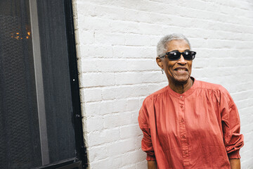 Obraz premium Senior black woman wearing sunglasses and smiling standing against a white brick wall