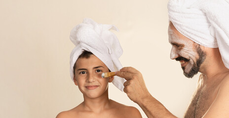 Boy with towel on his head smiles and looks at camera. Father applies mask with a brush. Father and...