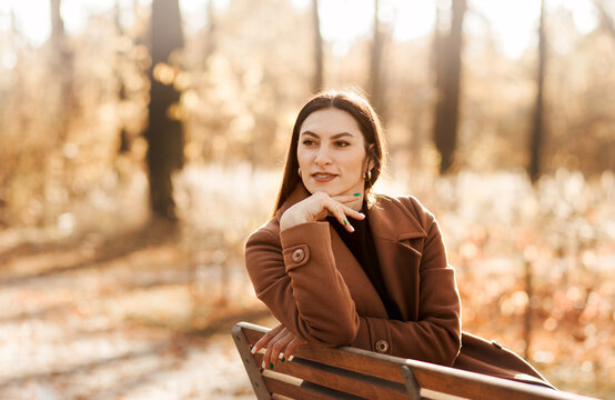 A young woman is a bruette sitting on a park bench in the fall in a coat