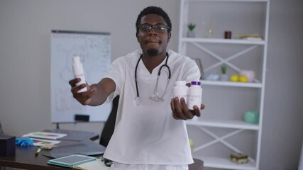 Rack focus confident African American doctor stretching vitamin bottles to camera posing indoors. Professional expert man advertising drugs in hospital with chemical formulas on whiteboard background