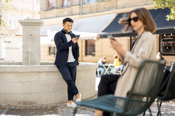 Young woman and asian man using smartphones on city area with fountain. Concept of city life. Beautiful girl wearing glasses sitting on shair. Guy with headphones. Warm sunny day