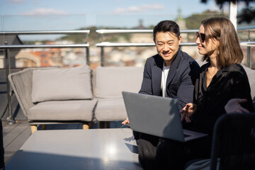 Businessman and businesswoman working with laptop on sofa on balcony. Smiling caucasian woman and asian man in formal wear. Concept of business cooperation. Sunny day. Blurred background of city