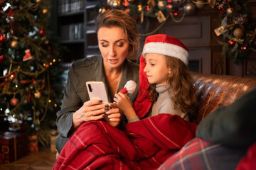 Mother and daughter in Santa hat looking at phone sitting in armchair in a Christmas decorated living room. Close view.