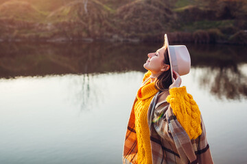 Woman relaxing by autumn lake at sunset. Stylish girl in hat enjoying fall landscape, breathing...