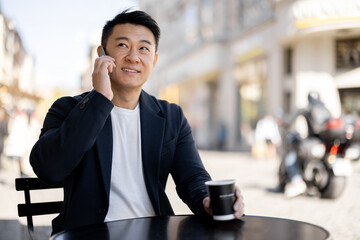 Asian businessman drinking coffee and talking on mobile phone at outdoor cafe. Concept of rest and leisure. Modern adult successful man wearing suit. Blurred background of sunny city street