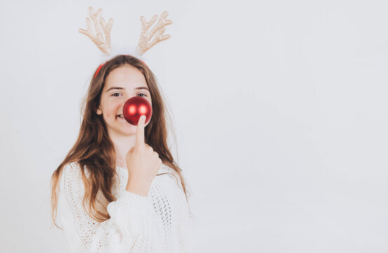 Young girl in white sweater and horns touches red bouble as nose christmas smile light