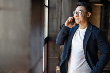 Smiling asian businessman talking on smartphone and looking from window in unknown building....