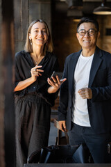 Smiling multiracial businesspeople look at camera during work in cafe. Concept of remote and freelance work. Teamwork and business cooperation. European woman with smartphone. Asian man with briefcase