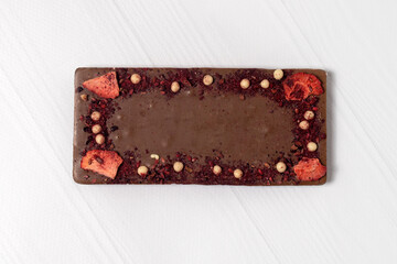 Craft homemade chocolate bar laying down on wooden white background, with pieces of nuts, fruits and berries. Exclusive dessert. Copy space.