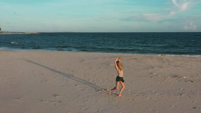 Young beautiful girl ballerina is dancing on the beach near the sea on the golden sunset. Moving woman, charming actress smile. Drone flies near dancer on the ocean shore. 