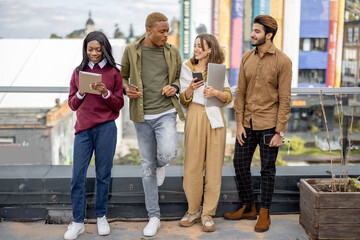 Multiracial students talking and using digital devices on balcony at university campus at day. Concept of education. Remote and e-learning. Idea of student lifestyle and friendship