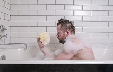 Sad man bathes in a bath with lush soap suds, he holds a sponge in his hands and looks thoughtfully.