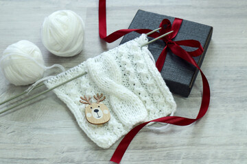 Preparing handmade gifts for Christmas: knitwear made of white yarn, balls of yarn, a deer figurine, a gift box tied with a red ribbon, side view.