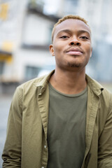Portrait of young black guy looking at camera outdoors. Handsome man wearing jacket and t-shirt. Blurred background of city