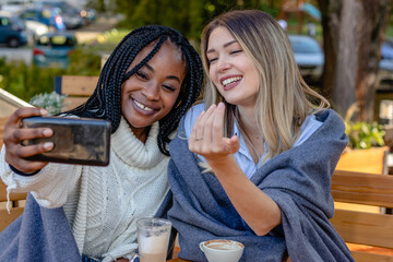 Girlfriends enjoy coffee together in a coffee shop, sitting at a table, laughing and bragging about engagement ring while taking a selfie with a smartphone. Diversity and multi-ethnic concept.