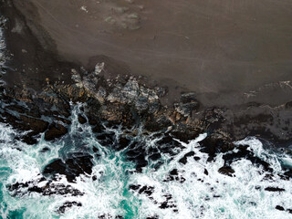 Overhead view of a beach with black sand and the blue water of the sea crashing against the rocks on the shore.