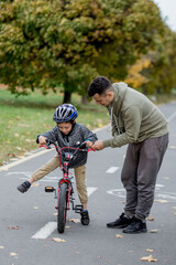 Father teaches his son to ride a bicycle on the bike path in the park. The father is holding a bicycle and the son is sitting on it