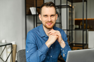 Fototapeta premium Smiling intelligent male freelancer using laptop for work online sitting at the desk and looks at camera in the coworking space, typing on the keyboard, writes article, chatting and messaging online