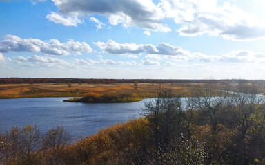 Beautiful autumn landscape in clear, sunny weather. Yellowed grass on the bank of a blue river under a bright, blue sky with clouds.
