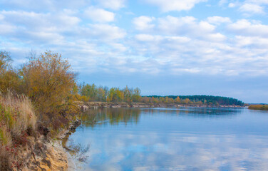 Green and yellow bushes on the steep bank of the river. Reflection of trees and clouds on the water surface of the river. Autumn landscape.
