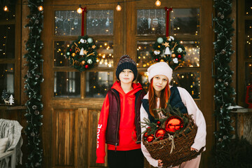 two children boy and teenage girl with stay on porch of village house with basket of toys and red balls, ready to decorate Christmas tree, during winter holidays, Christmas and New year vacation