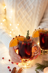 Young woman in knitted wool sweater is holding a glass with hot winter drink: mulled wine with spices, fruits. Christmas lights on background. Cozy atmosphere