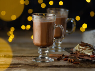 A beautiful composition - flavored cocoa in glass cups, pieces of chocolate, flickering yellow lights in the background. Coziness, Christmas spirit, romantic evening, Valentine's day.
