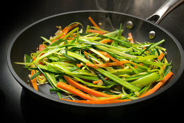 Steaming vegetable noodles from fresh carrot julienne and green leek strips in a black frying pan,...