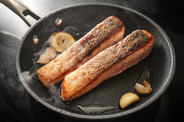 Two slices of fresh salmon fillet with a crispy fried skin in a black pan with garlic, thyme and...