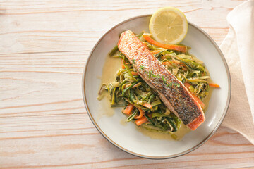 Fried salmon fillet with crispy skin on with leek and carrot vegetables on a plate, light wooden...