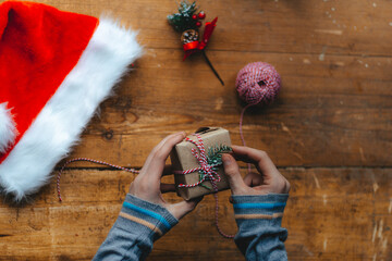 Christmas gift packaging. Hands wrap a small gift box in craft paper and a red-white string on the rustic wooden table with a Santa hat. Atmospheric image, Scandinavian nordic style