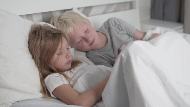 Children use their mobile phone in the morning in bed. The younger sister and brother play games on their phone at home.