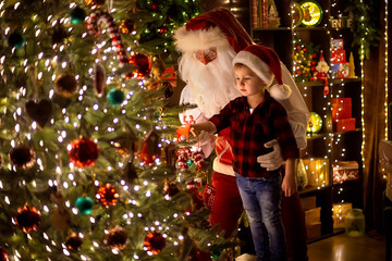 Santa Claus decorates Christmas tree with little boy assistant at home. New Year, Merry Christmas 
