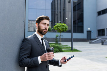 Bearded businessman in formal suit on break using mobile phone use smartphone. business man standing outside on modern urban city street background with coffee cup in downtown outdoors. copy space