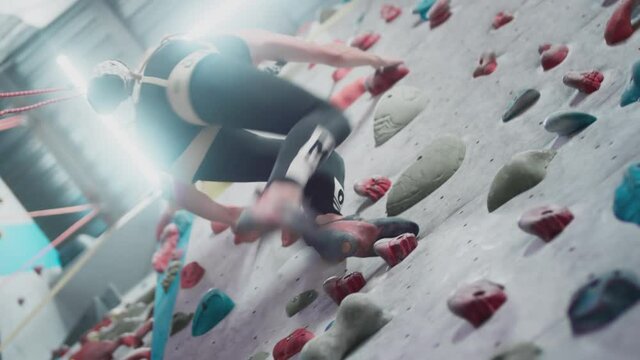 Sporty girl rock climber is engaged on an artificial wall in the gym. Athletic Woman Grasping stones and ledges. Climbs up using a safety rope and a carabiner. Bouldering in the city. Slow motion.