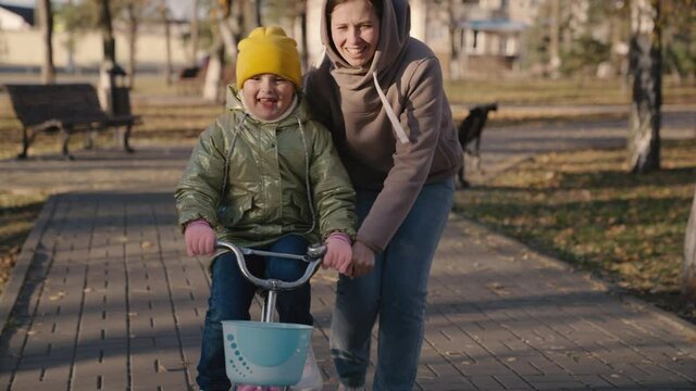 cheerful child learns ride two-wheeled bike with his mother, happy family, kid laughs pedals wheel while driving vehicle, baby play city park with his mother, parent helps girl learn move forward