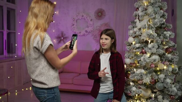 cute caucasian redhead single mother takes pictures of long-haired teenager son on phone against background of Christmas tree in living room decorated on New Year's Eve. Happy not complete family