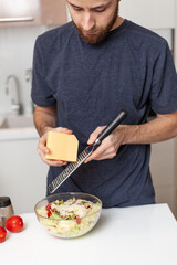 a young man with a beard 25-35 in a modern kitchen rubs cheese and prepares a salad