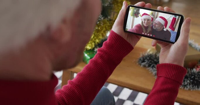 Caucasian man with santa hat using smartphone for christmas video call with smiling couple on screen