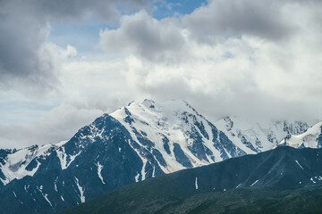 Awesome mountains landscape with high snowy mountain range among white clouds in blue sky. Atmospheric highland scenery with snow-white big mountain ridge in overcast weather. Wonderful snowy pinnacle