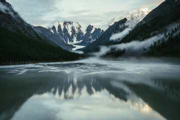 Naklejka na ściany i meble Scenic alpine landscape with snowy mountains in golden sunlight reflected on mirror mountain lake in fog among low clouds. Atmospheric highland scenery with low clouds on rocks and green mirror lake.